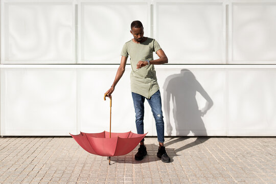 Serious African American male holding an upside-down umbrella on street on sunny day and checking time on wristwatch