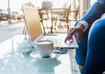 Side view of crop anonymous black businessman in formal suit sitting at table with cup of coffee and typing on laptop while working in cafe