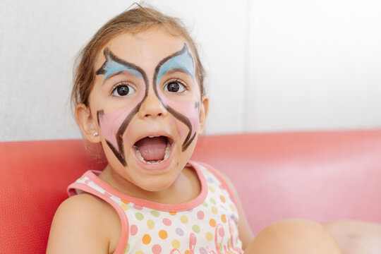Cheerful girl with picture of butterfly on face sitting on sofa at home while enjoying weekend and having fun