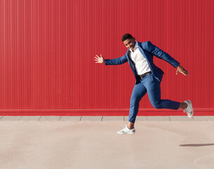 Full body side view of optimistic young African American businessman in stylish suit and sneakers running and jumping against red wall