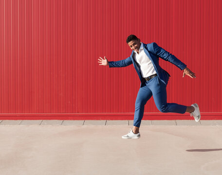 Full body side view of optimistic young African American businessman in stylish suit and sneakers running and jumping against red wall