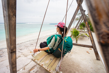 Side view of unrecognizable female traveler sitting on swings and enjoying fresh breeze while resting during summer holidays on sandy beach of Hulumale island in Maldives