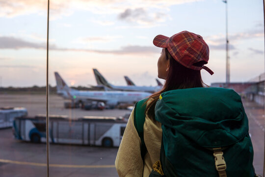 Back view of young Asian female traveler with backpack standing near window in airport terminal and observing airplanes while waiting for flight in Sri Lanka
