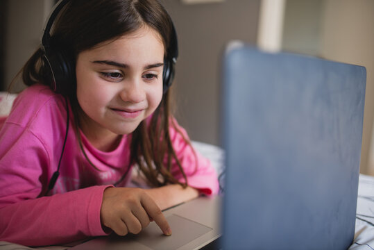 Focused little girl in headphones lying on bed and watching online video on laptop while spending free time at home