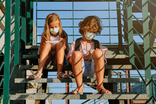 Two children with masks and school bags sitting on an industrial staircase doing their homework together on a sunny day