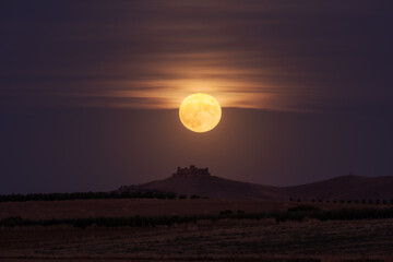 Bright yellow moon glowing on night sky over distant castle and fields in countryside