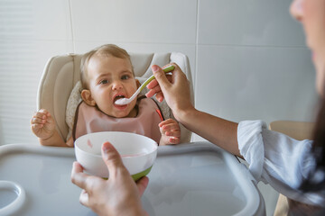 Little girl sitting on a high chair with a bib eating from the spoon with cropped unrecognizable mother feeding her