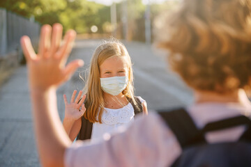 Two kids with school bag and mask greeting with her hand from a distance in the street in the morning