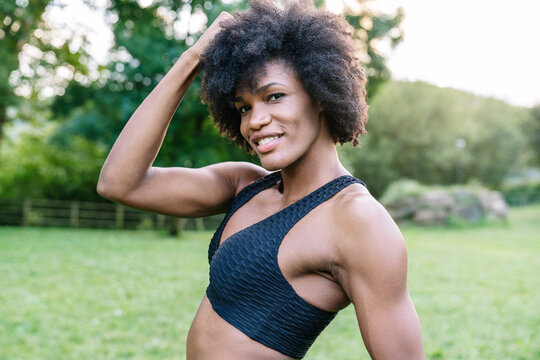 Young African American sportswoman with curly hair wearing black top looking at camera with smile while standing in green park during workout