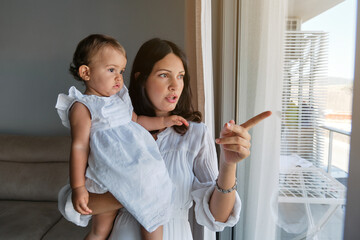 Mother with a little girl in white dress in her arms in front of a window pointing outwards in a house