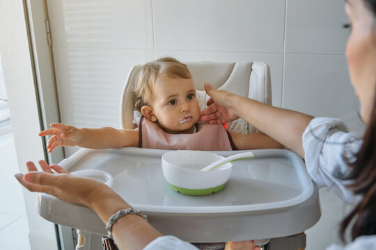 Unrecognizable mother removing the bib of a girl sitting in a highchair with the dirty mouth of food in the kitchen