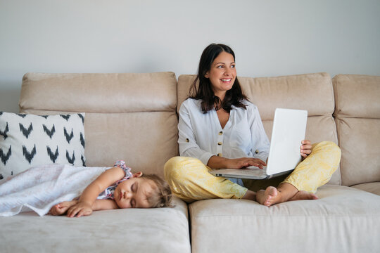 Woman working on the sofa with happy expression with the laptop while a toddler is sleeping on a side at home