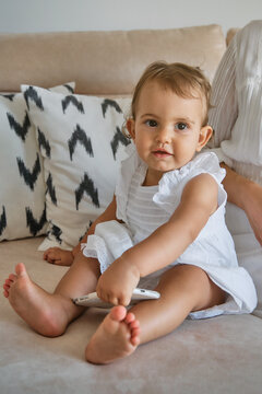 Vertical photo of a little girl in a white dress looking distracted while holding a mobile phone in her hands over a sofa