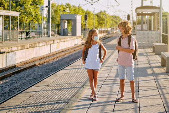 Two children with black school bags and mask waiting for the train on a platform at the station in the morning