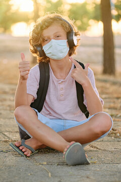 Vertical photo of a blond boy with mask and school bag sitting cross-legged while listening to music and making a gesture of being well by raising his thumb while facing the camera