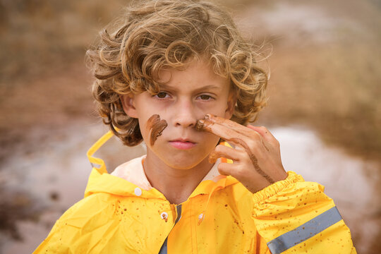 Blond boy with curly hair in a yellow raincoat drawing marks on his face with the mud looking at the camera seriously in the middle of the forest