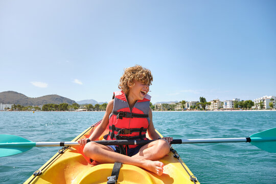 Blond boy with curly hair and a life jacket sitting in a yellow kayak in the sea with the beach in the background on a sunny day