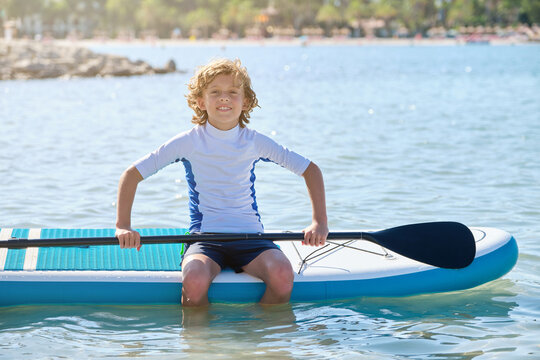 Blond teenager with curly hair sitting on a paddle surfboard in the middle of the water with the paddle in his hands facing the camera
