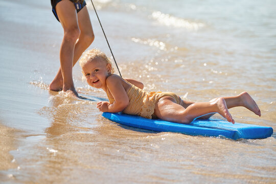 Little girl with a swimsuit and headband facing the camera while lying on a surfboard at the edge of the beach playing while someone drags her along the waves
