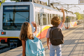 Two children with black school bags on their backs greeting a train as it leaves the track from a station platform