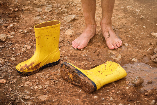 Dirty muddy bare feet with a pair of water boots next to them on a muddy road on a rainy day