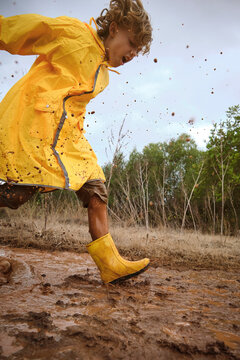 Vertical photo of a child in a raincoat and yellow boots playing into a puddle splashing water in the middle of a path in the forest