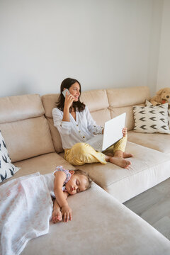 Little girl sleeping on the sofa while her mother works with a laptop and talking with the mobile phone next to her
