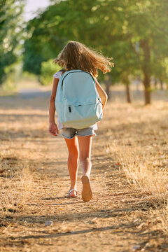 Vertical photo of the portrait of the back of a blond girl with a blue school bag walking along a tree path