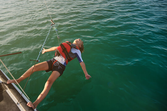 Blond boy with life jacket hooked by a rope with his feet on the edge of a boat doing a freedom gesture while sailing in the middle of the sea