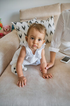 Vertical photo of a little girl in a white dress crawling on a sofa with her mother and a mobile phone next to her at home