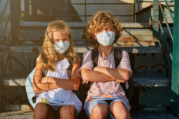 Two blond kids with masks and school bags sitting on an outside staircase with their arms crossed in denial while facing the camera