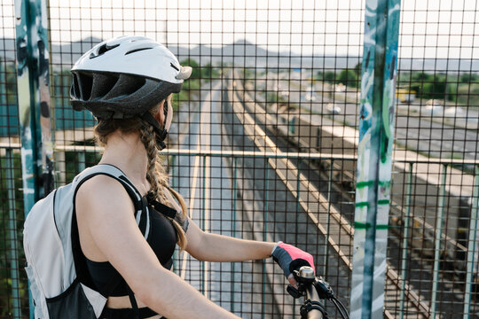 Back view of crop unrecognizable female bicyclist in protective helmet and gloves standing near grate fence on bridge over road with blurred hills in background