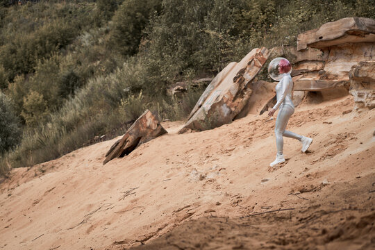 Positive young female with dyed red hair wearing silver space suit and glass helmet and looking away while walking on rocky formation