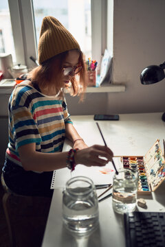 High angle side view of concentrated female artist standing at table in modern workshop and using watercolor for painting