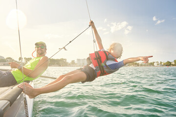 Blond boy with a life jacket attached to a rope from a boat hanging out to sea pointing to the horizon with a man next to him sitting on the boat steering the wheel with the sun illuminating them