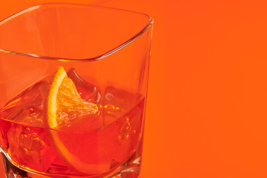 Crystal glass of alcohol drink with slice of lemon and ice cubes placed on table in studio on orange background