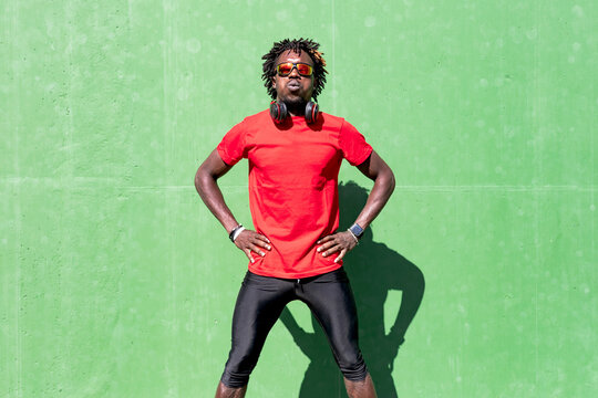 Tired African American male athlete in sportswear and with headphones relaxing on street after training on sunny day while exhaling and looking at camera