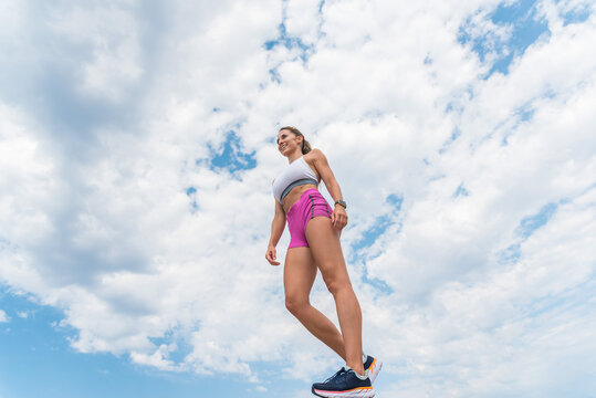 From below of smiling fit female athlete in sportswear standing on background of blue cloudy sky and looking away while relaxing after training