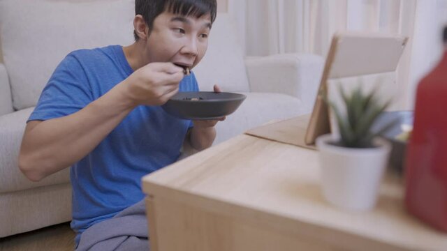 Asian young man enjoys watching a tablet while eating a spoon of riceberry in cozy living room, people and daily routine, having a meal, nutrition and delicious recipe, self entertainment at home 