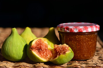 Fresh green figs and homemade fig jam in a glass, on a wicker table, against a dark background
