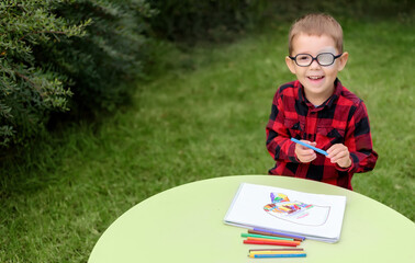 A little boy drawing a picture in the garden (outdoors). Wearing glasses and an eye patch (plaster,...
