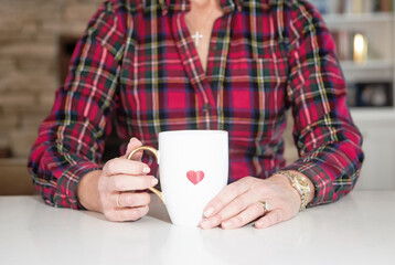 Original faceless portrait of a woman dressed in red tartan plaid sitting a table holding a white coffee cup in her hands with a red heart