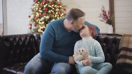 Positive father kissing little daughter and combing girl's hair on Christmas eve. Portrait of happy man and child spending New Year at home. Joy and celebration concept. - Powered by Adobe