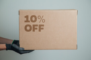 Detail of man with black gloves holding a cardboard box with 10% discount for delivering products on gray background. Delivery concept. Delivery service concept. Copy space.