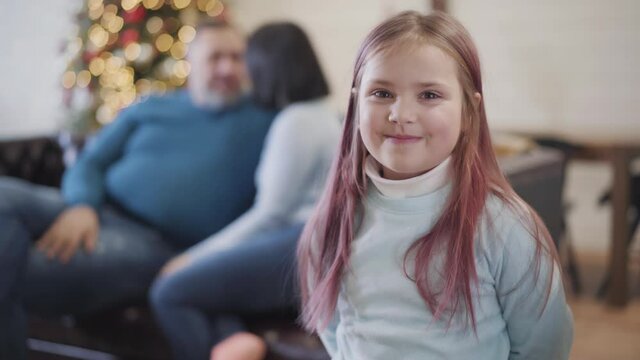 Cheerful Caucasian daughter looking at camera and running back to happy parents sitting on couch at the background. Charming girl enjoying Christmas holidays celebration at home. Rack focus.