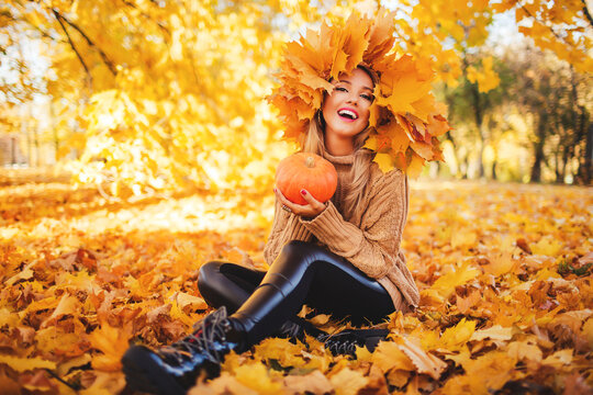 Outdoors lifestyle fashion image of happy beautiful girl sitting on the grass in autumn park. Holding a pumpkin. Wearing stylish knitted pullover and wreath of maple leaves. Autumn colors. Happiness 

