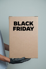 Detail of man with black gloves holding a cardboard box written black friday to deliver products on gray background. Delivery concept. Delivery service concept. Copy space. Black friday concept.