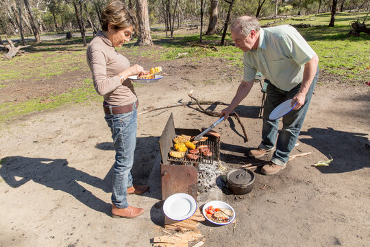 Grey nomad couple tending food cooking on grill at campfire