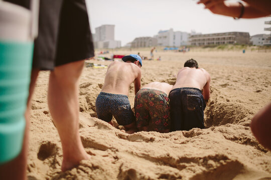 A group of youthful guys bending over and digging a hole in the sand.