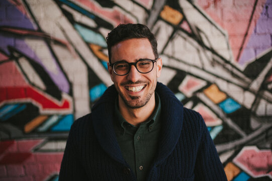 Young man wearing glasses and smiling in front of graffiti brick wall